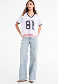 Hvid mesh-jersey med marineblå kant og "81" print, lagd over en lyserød top. Lys blå vide ben-jeans med en afslappet pasform og sneakers.