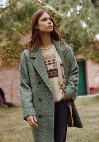Manteau en laine vert à motif chevrons avec un design double boutonnage, porté sur un pull crème à motifs et un jean foncé, sur fond naturel.