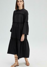 Zwarte lange mouw tiered jurk van zachte stof, met ruchesdetails aan de naden. Gepaard met tan kleurige sandaaltjes met bandjes.
