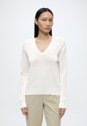 Donna con capelli lisci neri indossa un maglione bianco con scollo a V e pantaloni beige su uno sfondo bianco semplice.