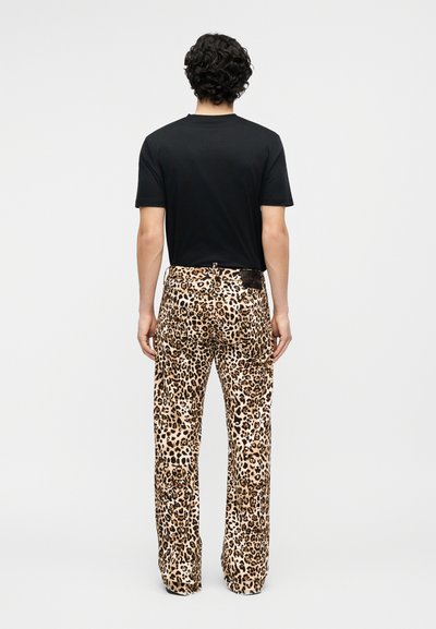 Άτομο με σγουρά μαλλιά που κοιτάζει προς τα πίσω, φορώντας απλό μαύρο μπλουζάκι και ίσια παντελόνια με λεοπάρ τύπωμα σε λευκό φόντο.