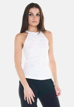 Jeune femme aux longs cheveux bruns portant un haut blanc sans manches froncé et un pantalon noir, debout devant un fond clair uni.