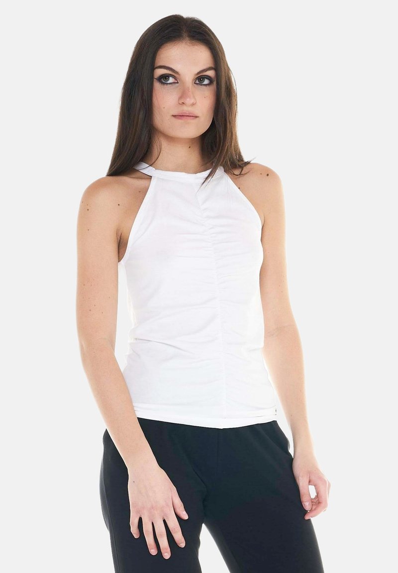 Jeune femme aux longs cheveux bruns portant un haut blanc sans manches froncé et un pantalon noir, debout devant un fond clair uni.