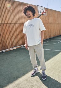 Nike-logo t-shirt i cream, parret med lysegrå sweatpants med sidelommer. Modellen har grå sportssko på en bane.
