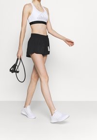 Sostén deportivo blanco con logo negro, pantalones cortos negros con cinturilla elástica y zapatillas deportivas blancas. La modelo lleva una cuerda de saltar negra.