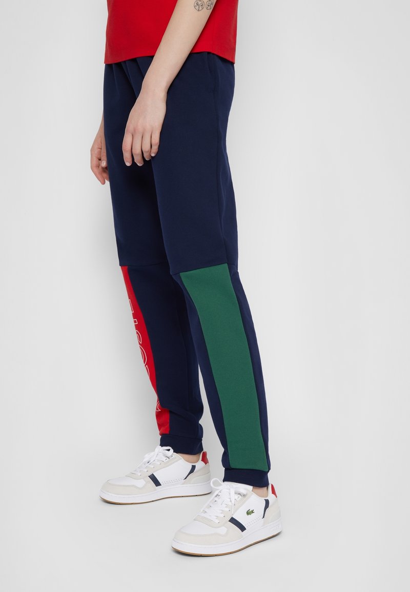 Marina joggers med röda och gröna sidopaneler, med vitt logotext. Bärs med vita sneakers, vilket framhäver den sportiga, avslappnade designen.