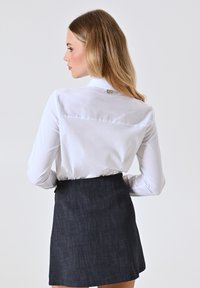 Blusa bianca a maniche lunghe con chiusura sul retro, abbinata a una gonna strutturata di colore blu scuro. L'outfit presenta una silhouette aderente.