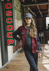 Bordeaux varsityjack met zwarte mouwen en witte strepen, gecombineerd met een donkergrijze geblokte shirt en zwarte wijde broek. Grijze pet gedragen.