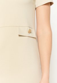 Beiges Kleid mit strukturiertem Obermaterial, kurzen Ärmeln und einer Seitentasche mit einem dekorativen Knopf.