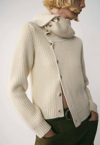 Creme strik cardigan med en høj, voluminøs krave, der har knaplukninger og ribbede manchetter, parret med mørkegrønne bukser.