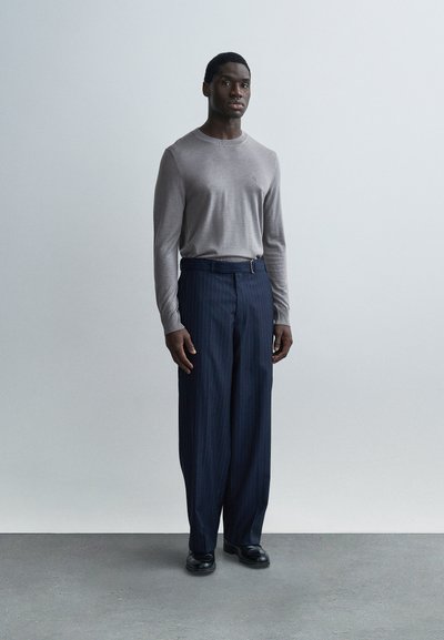 Pull gris à manches longues associé à un pantalon bleu marine à fines rayures. Le pantalon présente une texture lisse et un détail ceinture. Des chaussures noires complètent la tenue.