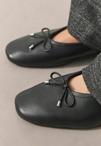 Bailarinas de cuero negro con punta redondeada, decoradas con un lazo y terminales plateados en los cordones. Parte superior texturizada, acabado suave.