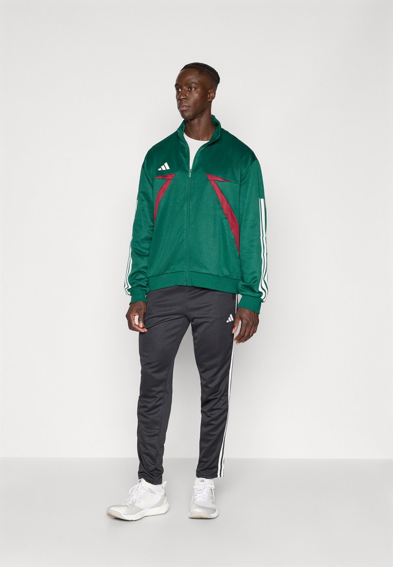 Groene ritsjack met bordeauxrode accenten, witte strepen op de mouwen; gecombineerd met zwarte broek met witte zijstrepen en lichte sportschoenen.