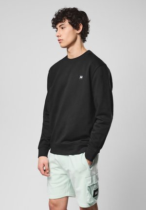 Młody mężczyzna z kręconymi włosami, ubrany w czarną bluzę i jasne krótkie spodenki typu cargo, na jednolitym tle.