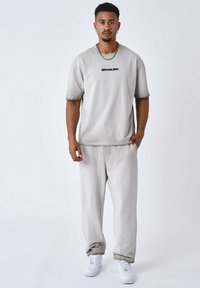 Camiseta oversized gris claro con un logo negro, combinada con pantalones de corte relajado gris claro y zapatillas blancas, de pie sobre un fondo neutro.