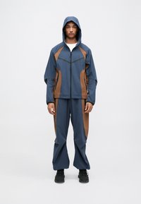 Μπλε και καφέ σετ δύο κομματιών με τζάκετ που κλείνει με φερμουάρ και διαθέτει κουκούλα, αντίθετα πάνελ και χαλαρούς παντελονιού, κατασκευασμένο από ελαφρύ ύφασμα.