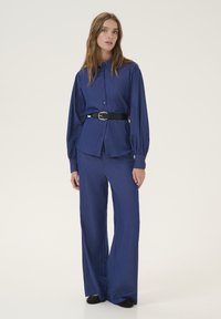 Modrý dvoudílný outfit skládající se z košile na knoflíky s mírně oversize střihem a kalhot s širokými nohavicemi, doplněný černým opaskem.