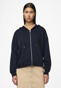Felpa zip con cappuccio in cotone color navy. Presenta un cappuccio con cordino, tasche frontali e polsini a costine, abbinata a una gonna beige con bottoni.