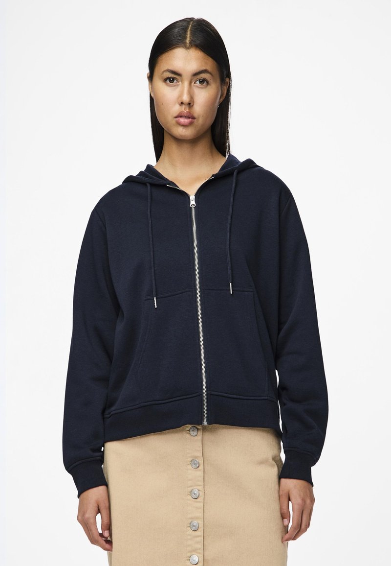 Felpa zip con cappuccio in cotone color navy. Presenta un cappuccio con cordino, tasche frontali e polsini a costine, abbinata a una gonna beige con bottoni.