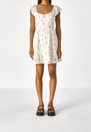 Vit blommig klänning med korta volangärmar, fyrkantig halsringning och samlad midja. Har små rosa blommor och en flerfaldig fåll.