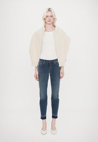 Gaišbeige oversize jaka, pieguļoša balta blūze un tumši džinsu bikses. Modelis valkā krēmkrāsas norādītas kurpes. Vienkāršs dizains, gluda tekstūra.