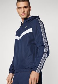Giacca sportiva navy con strisce bianche sul petto e lungo le maniche; presenta chiusura a zip frontale, cappuccio e polsini e fondo elastici.