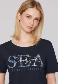Camiseta de manga corta en azul marino, hecha de una tela suave, que presenta un estampado gráfico con "SEA Soccer Spirit Sunset Sailing" en varias tonalidades de azul.
