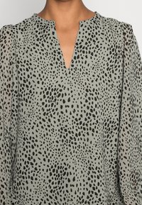 Ljusgrön blus med V-ringning, med ett svart prickigt mönster och genomskinliga puffärmar, tillverkad av ett slätt tyg.