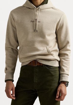 Beżowa bluza z kapturem z przednią kieszenią i ściągaczami, haftowana "Polo Ralph Lauren,", noszona z ciemnozielonymi spodniami i brązowym skórzanym paskiem.