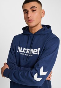 Granatowy hoodie wykonany z tkaniny o miękkiej strukturze. Posiada biały logo oraz akcenty na rękawach, w tym wzór w zygzak.