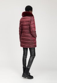 Persona vistiendo un abrigo acolchado burdeos con cuello de piel, pantalones de cuero negros y botas negras de tobillo, de pie contra un fondo simple.