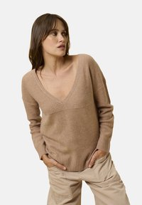 Beiger Strickpullover mit tiefem V-Ausschnitt, gerippten Bündchen und Saum. Weiche Textur, lockere Passform, kombiniert mit hellen Cargo-Hosen.