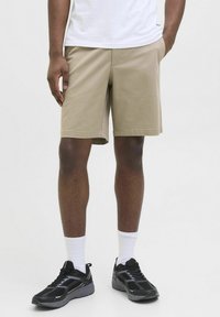Pantaloni scurți din bumbac bej deschis, cu o croială dreaptă și buzunare laterale, purtați cu un tricou alb simplu și pantofi sport negri.