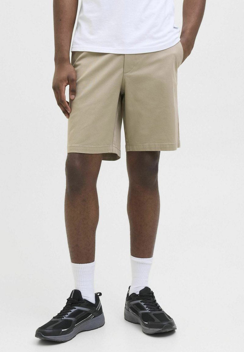 Pantaloni scurți din bumbac bej deschis, cu o croială dreaptă și buzunare laterale, purtați cu un tricou alb simplu și pantofi sport negri.