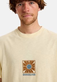 Világos sárga, rövid ujjú póló, puha anyagból készült. Jellemzője egy négyzet alakú grafika barna és kék napsugár designnal, amelyen a "QUIKSILVER" felirat található.
