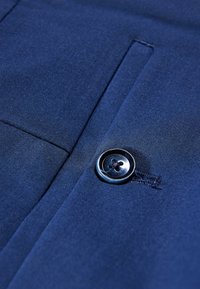 Nahaufnahme eines blauen Stoffs mit einem Knopf an einem Kleidungsstück, wobei eine Taschennaht und eine feine Textur sichtbar sind, was auf hochwertiges Material hinweist.