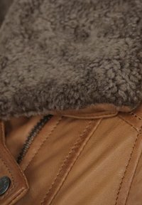 Brun læderjakke med en brun, fluffy shearlingkrave. Har synlige sømme og en sølvlynlås, der fremhæver de teksturerede materialer.