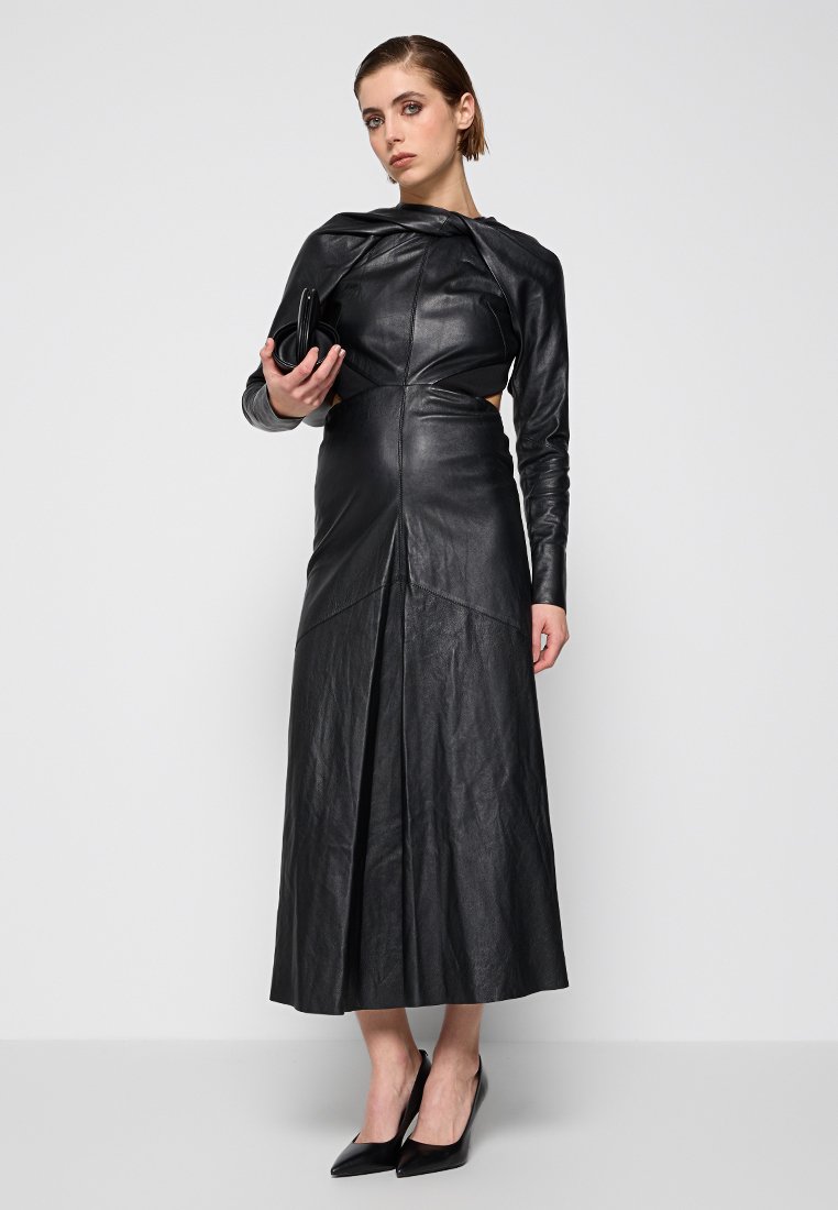 Czarna skórzana sukienka z długimi rękawami, dopasowanym gorsetem i plisowaną pełną spódnicą. Modelka trzyma małą czarną torebkę i nosi szpiczaste czarne szpilki.