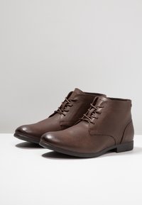 Botas de tobillo de cuero marrón con un acabado suave, punta redonda y diseño de cordones. Presentan un tacón bajo y detalles de costura mínimos.