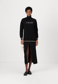 Zwarte coltrui met logo, gecombineerd met een zwarte splitrok met contrasterende stiksels. Het model houdt een kleine witte handtas vast.
