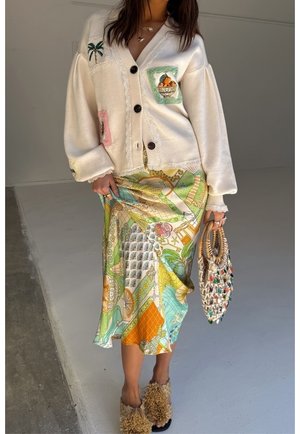 Vrouw die een crèmekleurige cardigan draagt met fruit- en palmboompatches, een kleurrijke patroonrok, textuurrijke slip-on schoenen, en een geweven tas in haar hand houdt.