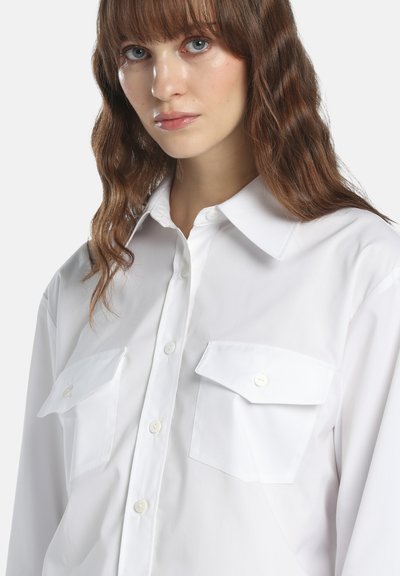 Jeune femme aux cheveux bruns ondulés portant une chemise blanche boutonnée avec deux poches à rabat sur la poitrine, sur un fond clair uni.