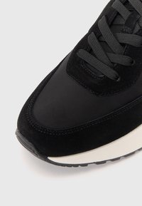 Črne športne copate, ki združujejo gladke tekstilne in usnjene materiale. Zaobljen nos, teksturirane vezalke in kontrastna bela srednja podloga.