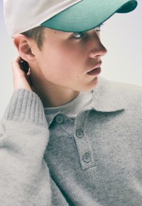 Szürke kötött pulóver galléros dizájnnal és háromgombos elülső résszel, világos ing felett viselve, fehér és zöld sapkával párosítva.