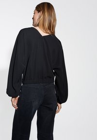 Blusa nera con retro a V, maniche lunghe a sbuffo e vita elastica, abbinata a jeans in denim scuro con tasche posteriori.