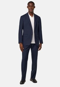 Costume bleu marine avec un design ajusté, blazer à deux boutons, revers crantés et pantalon assorti. Porté par-dessus une chemise polo blanche et des chaussures noires.
