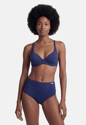 Woman with natural afro hairstyle wearing navy blue bra and high-waisted briefs, standing with arms relaxed against white background.