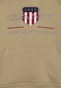 Felpa beige con cappuccio caratterizzata da un logo scudetto ricamato con strisce rosse e bianche e il testo "GANT AMERICAN SPORTSWEAR" in bianco sottostante. Tasche a marsupio.