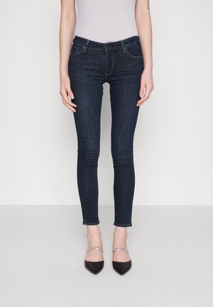 Wąskie, ciemnoniebieskie jeansy z denimu noszone przez osobę stojącą, zestawione z czarnymi spiczastymi butami z srebrnymi paskami na jednolitym tle.