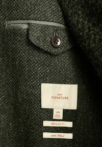 Dunkelgrüne Wolljacke aus strukturiertem Stoff, mit einem einzelnen Knopf am Kragen und Innenetiketten, die Größe und Pflegehinweise anzeigen.